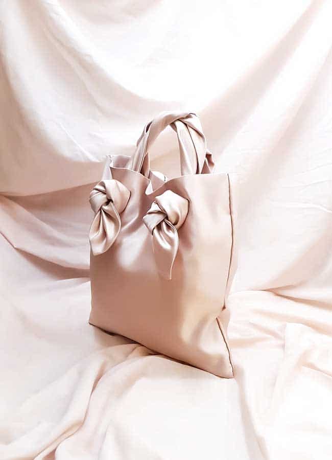 A blush pink satin tote with knot details on the straps by August NIght sits on a pink sheet which is draped rising up behind it too