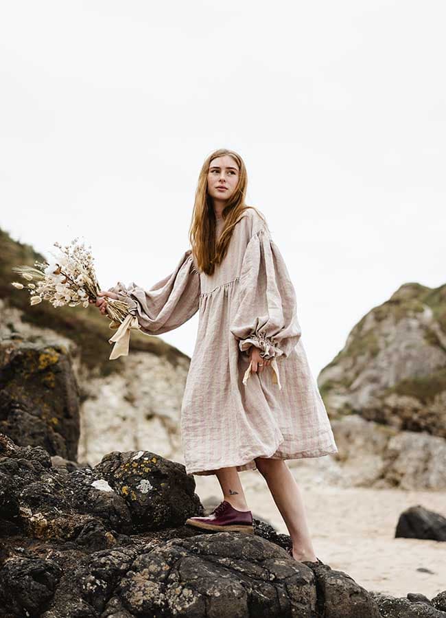 A model climbs rocks on a beach wearing a linen dress with dramatic voluminous sleeves from the SS21 collection of Northen Irish Kindred of Ireland carrying a bunch of flowers