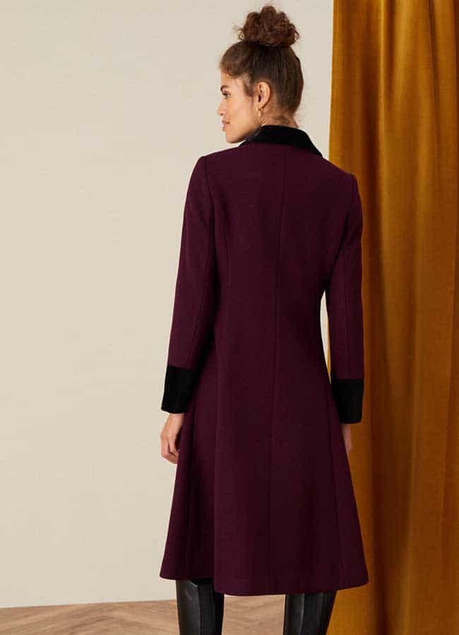 A model stands against a cream wall beside a wooden wall wearing a maroon coat with black cuffs and collar and a pair of black trousers