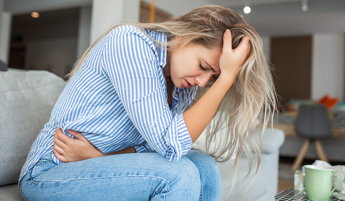 Woman in painful expression holding hands against belly suffering menstrual period pain, lying sad on home bed, having tummy cramp in female health concept