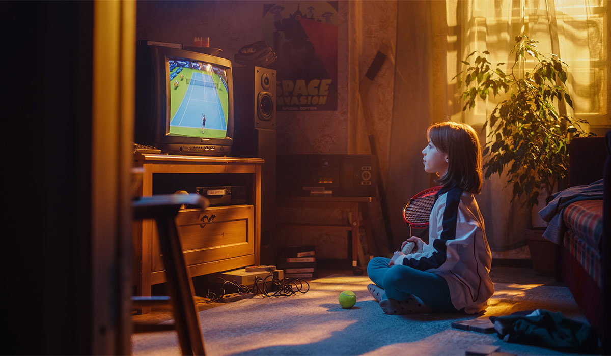 Young Sports Fan Watches a Tennis Match on TV at Vintage Home. Curious Girl Supporting Her Favorite Player, Imitating the Game with Her Racket. Nostalgic and Retro Childhood Concept.